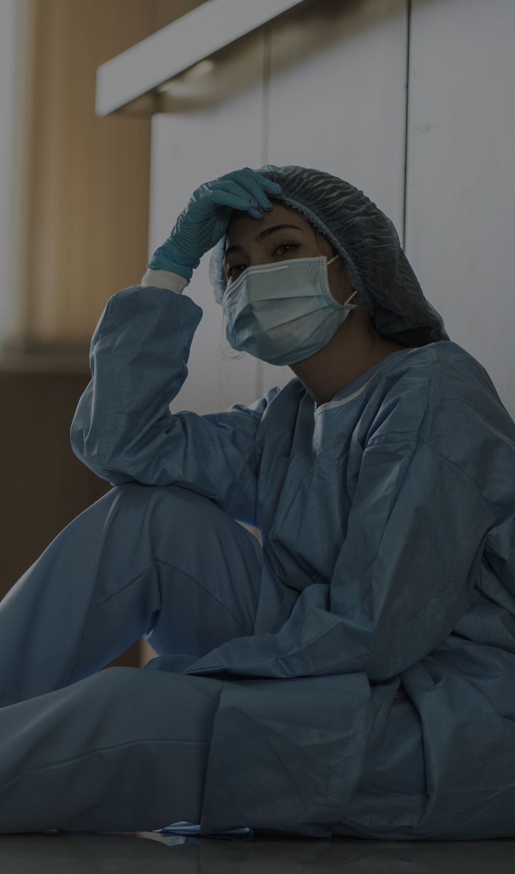 A healthcare worker sits on the floor in full surgery scrubs with a face mask.