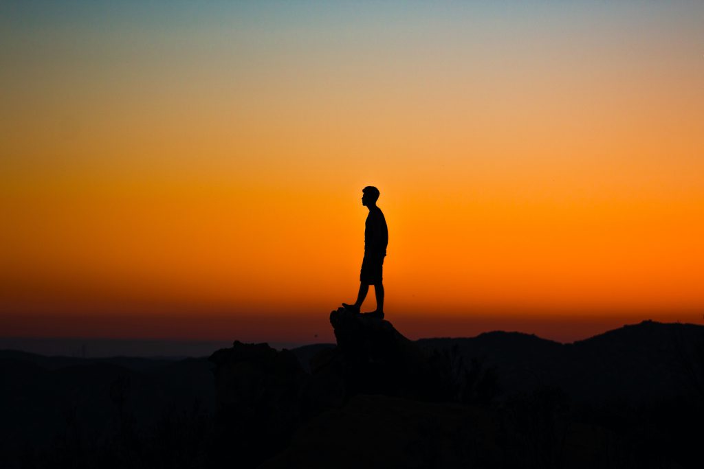 Silhouette of a man standing on an overlook with an orange fading sky in the background.