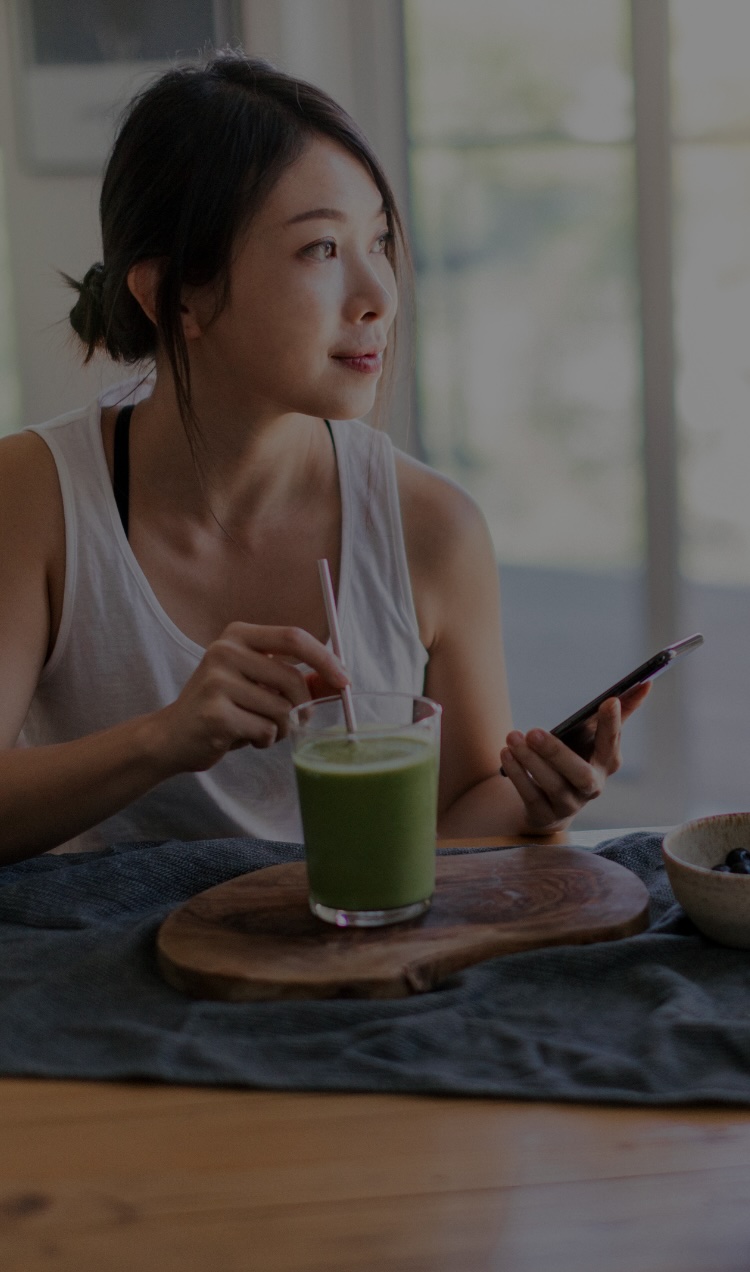 A woman sits at the kitchen table with a smoothie while on her phone.