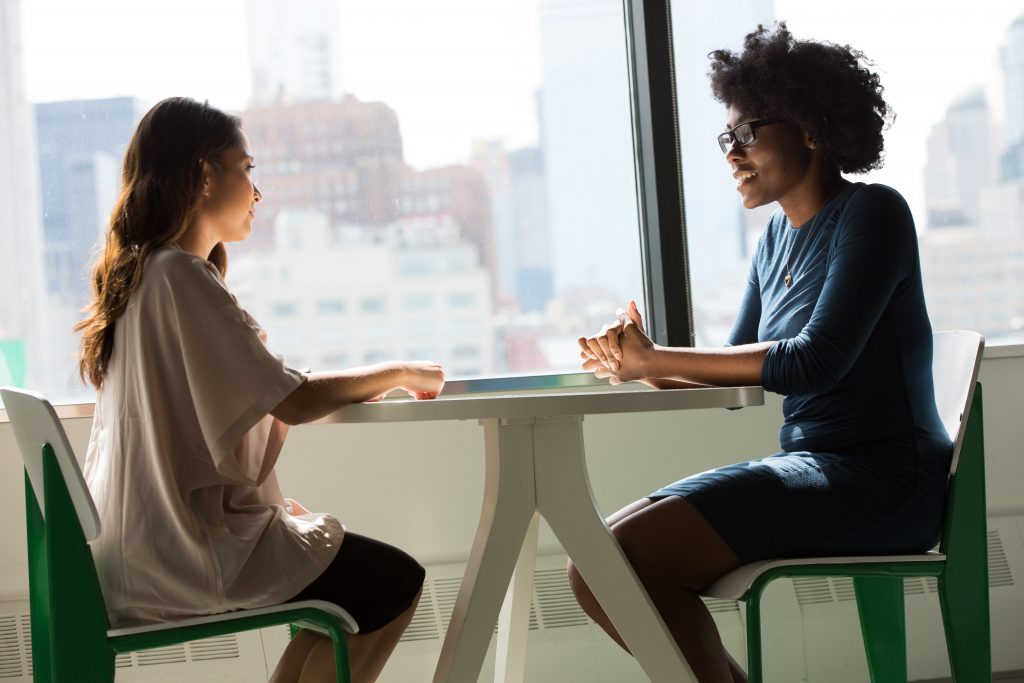 Two women sit in front of a large window at a small table, talking politely.