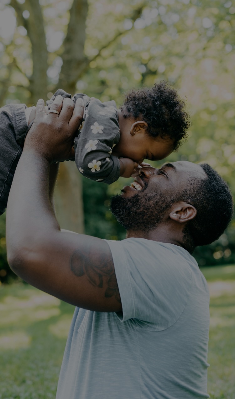 A dad holds his baby in the air with their faces nose to nose, both giggling and smiling.