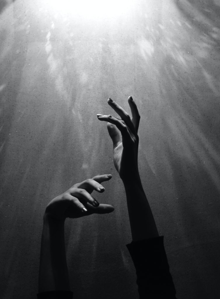 A black and white photo of someone reaching up towards a light with both hands.