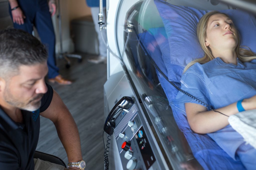 An HBOT technician sits next to a woman receiving treatment in an oxygen chamber.