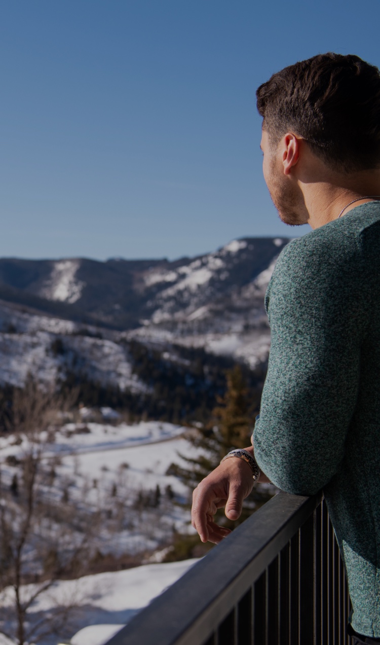 A man looks out on the snowy mountains as he leans on the rail of a balcony.
