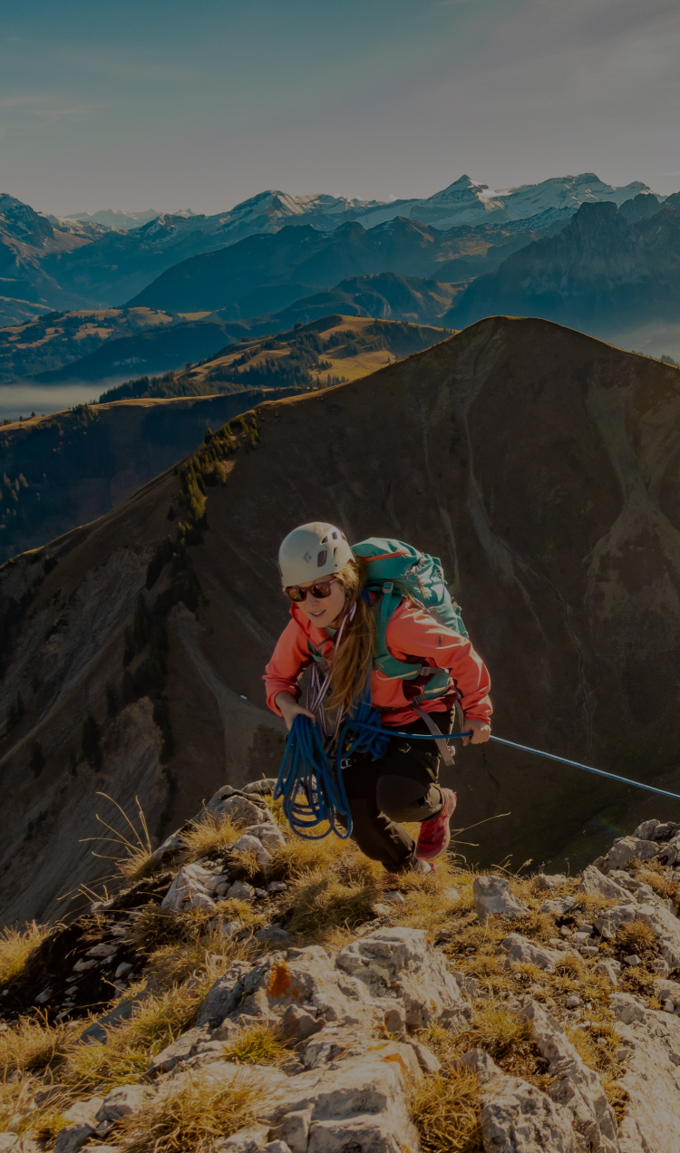 A climber reaching the top of a summit with a mountain range scraping the clouds behind her.