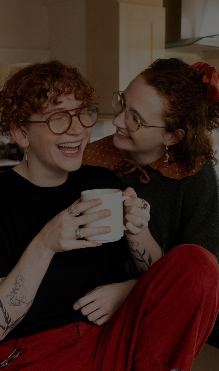 A queer couple smiles and laughs together over coffee.