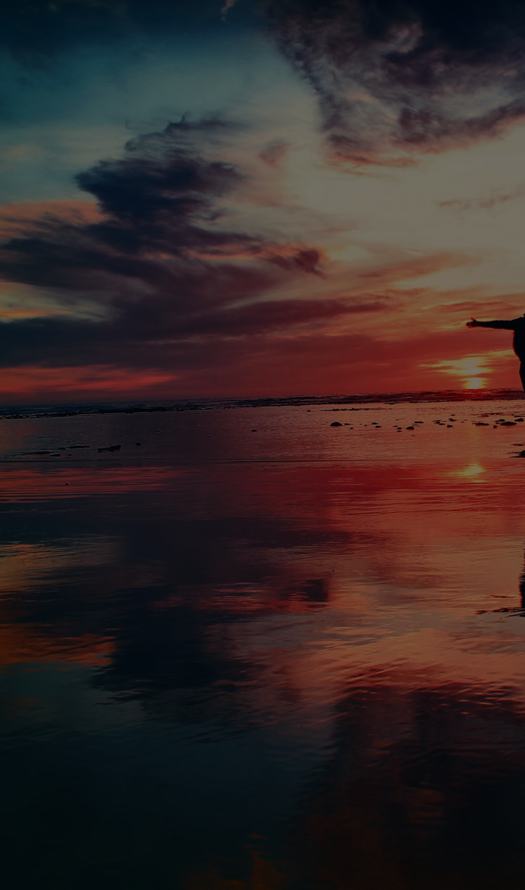 Silhouette of a person standing on the beach with their arms outstretched during a rainbow sunset.
