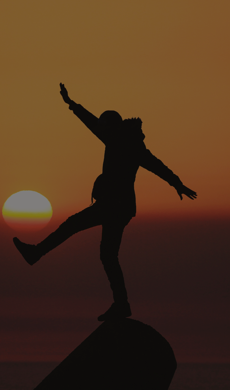 A person balances on the peak of a mountaintop at sunset.