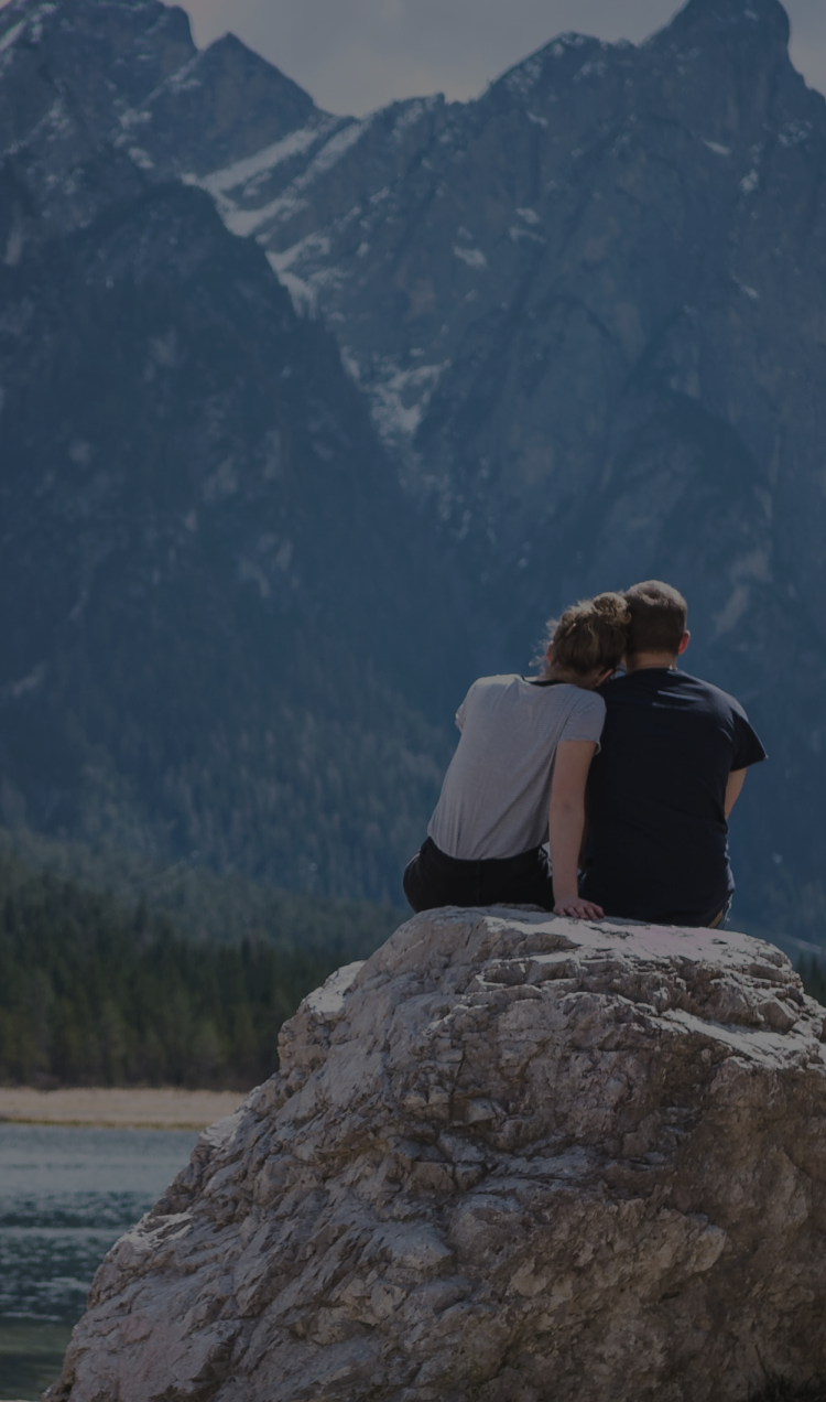 A couple sits on a rock overlooking a lake with a mountain landscape.