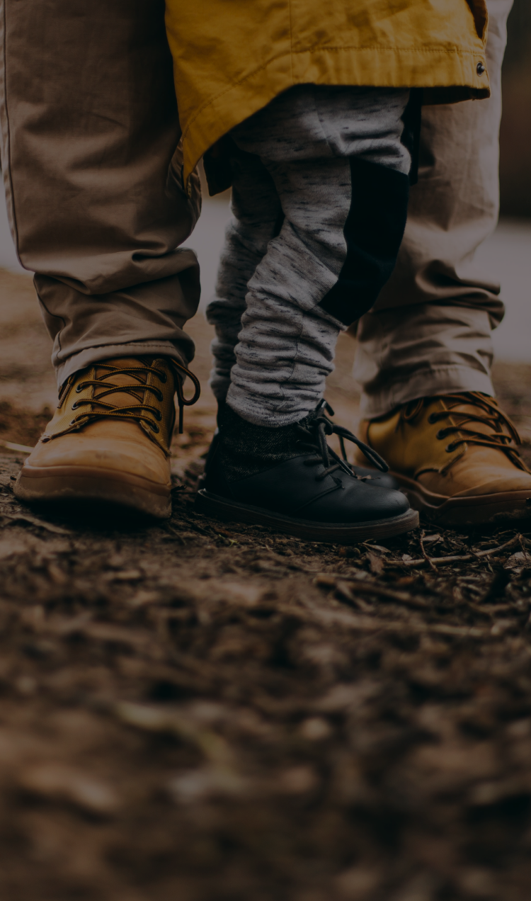 A parent stands with their toddler standing between their feet.