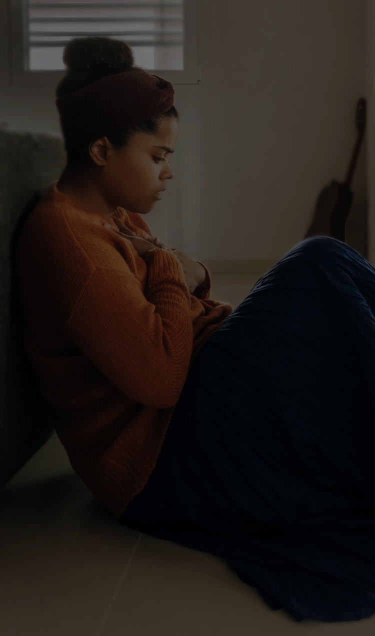 A woman sits on the floor at the foot of her bed with her hand on her heart.