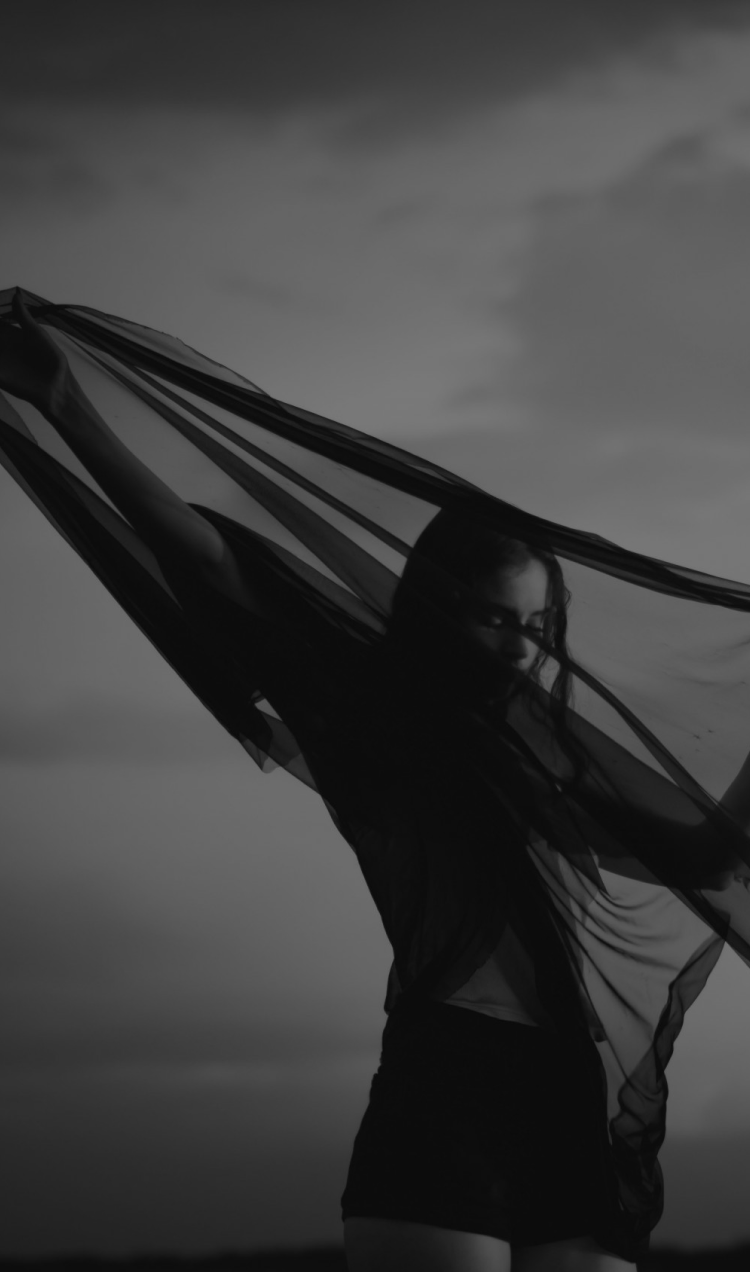 A woman stands draped in a black veil with the sky behind her.