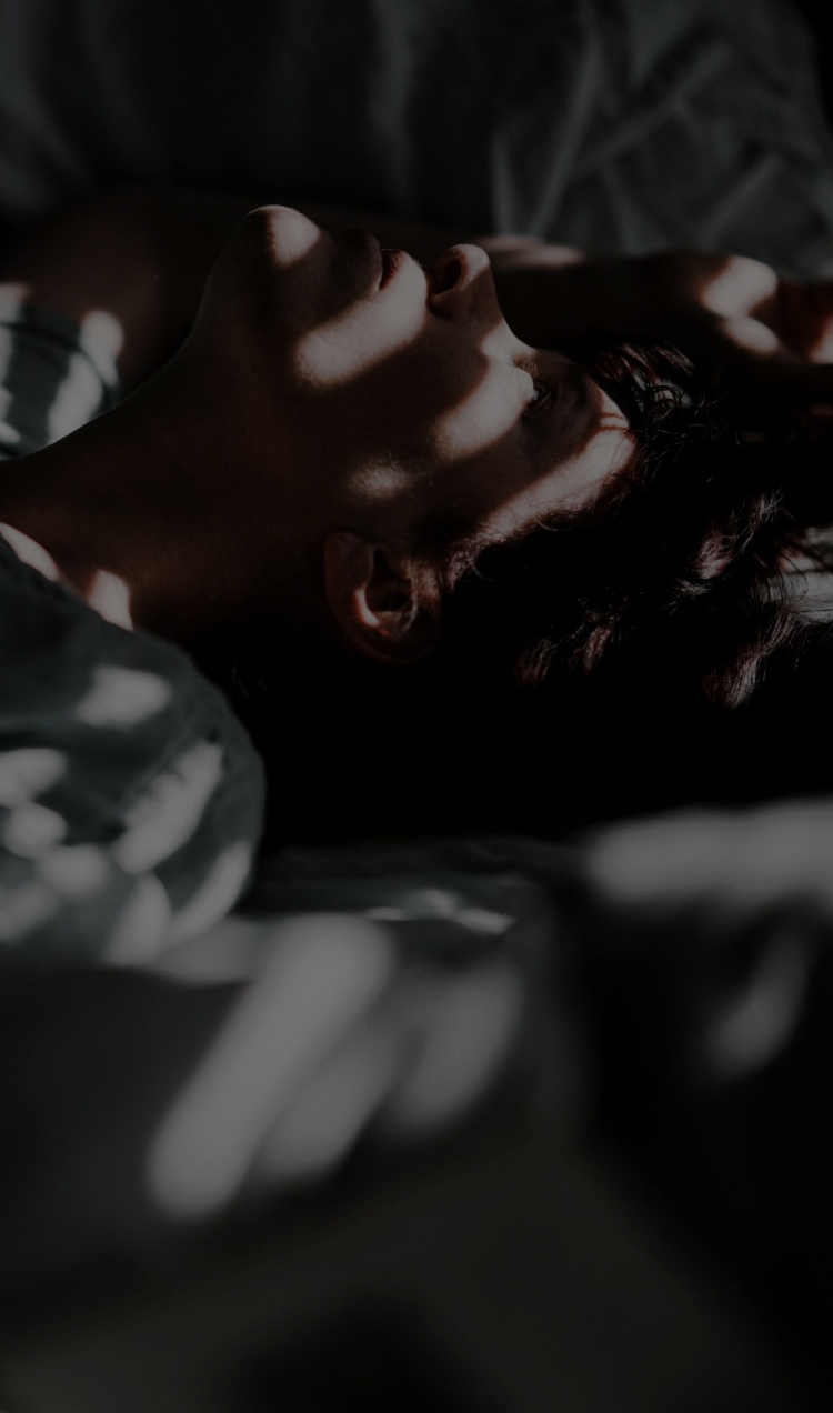 Someone laying in bed face up with light filtering through the blinds.