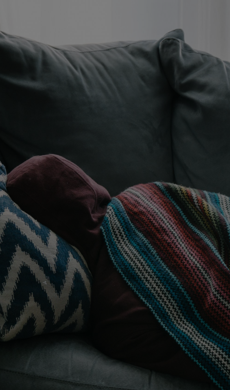 Someone bundled in a hoodie and blanket laying on a couch facing away from the camera.