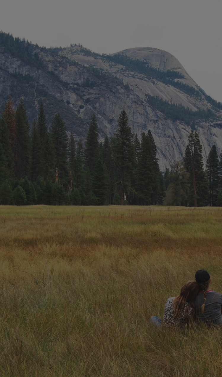 A couple sits in a field surrounded by mountains.