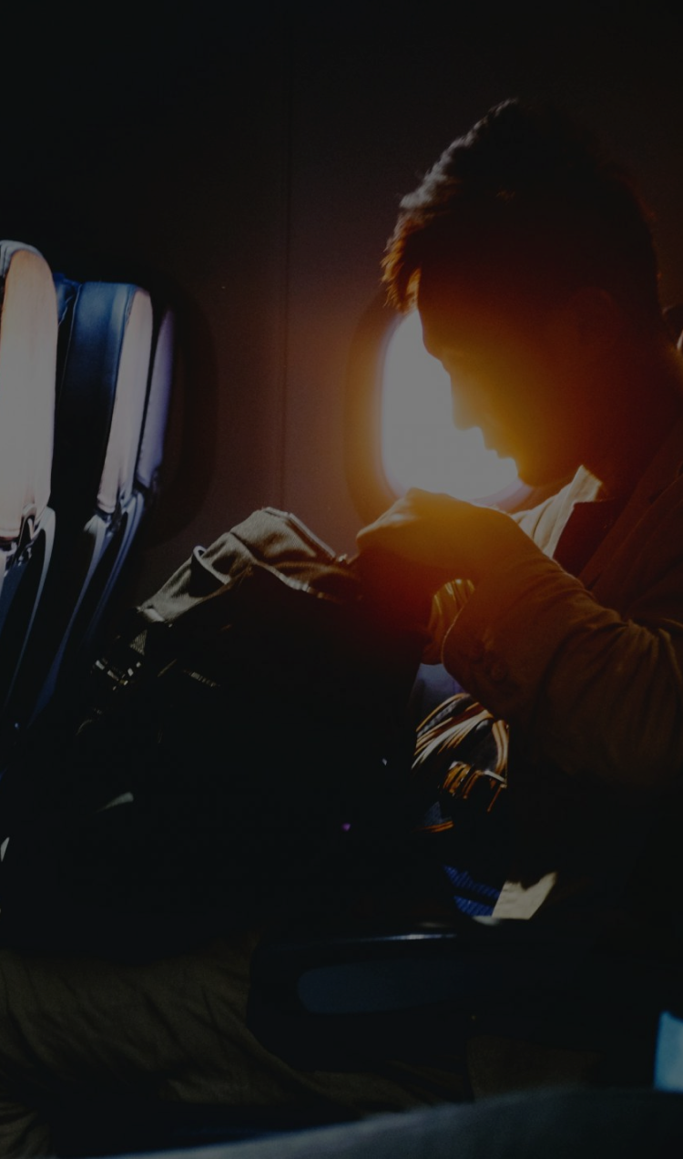 A man sits in an airplane with a travel packpack on his lap as the sun sets out the window.