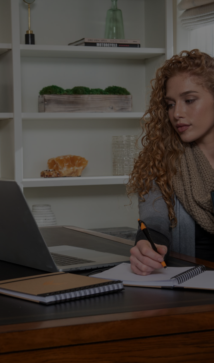 A woman sits at a desk taking notes while she attends virtual therapy.
