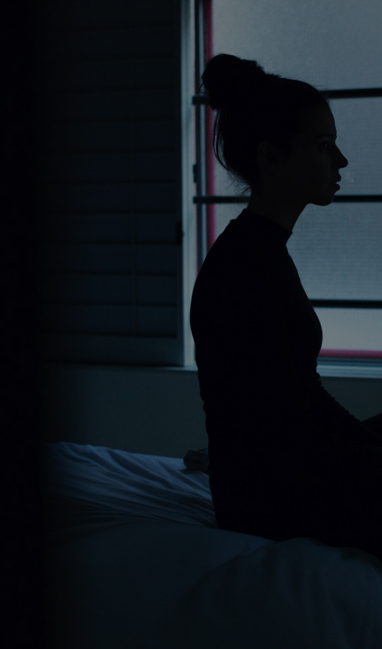 A woman sitting on the edge of a bed in a dimly lit room, backlit by a window.