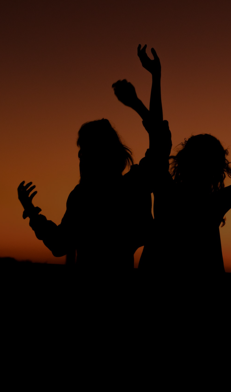 Three women with their arms raised, backlit by an orange sunset.