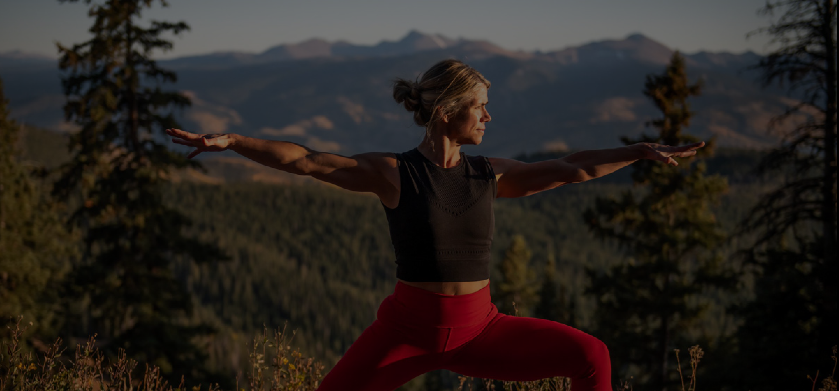 A woman in a black crop top and red leggings does warrior pose outside with the mountains behind her.