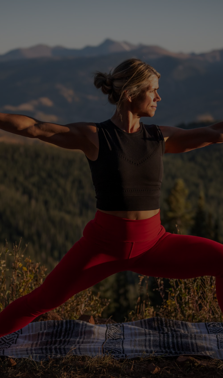A woman in a black crop top and red leggings does warrior pose outside with the mountains behind her.